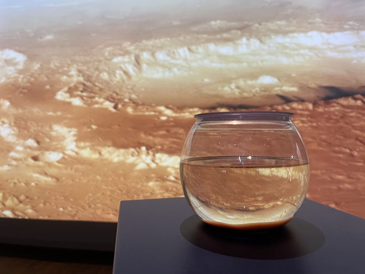 Glasbehälter mit Wasser vor einer warmfarbigen Projektion der Marsoberfläche.