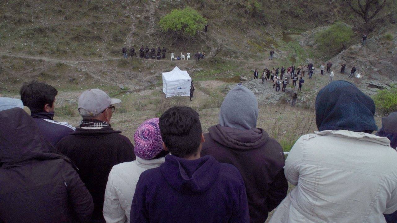 People are gathered on a hillside, watching an event near a tent, with several individuals and a group in the background.