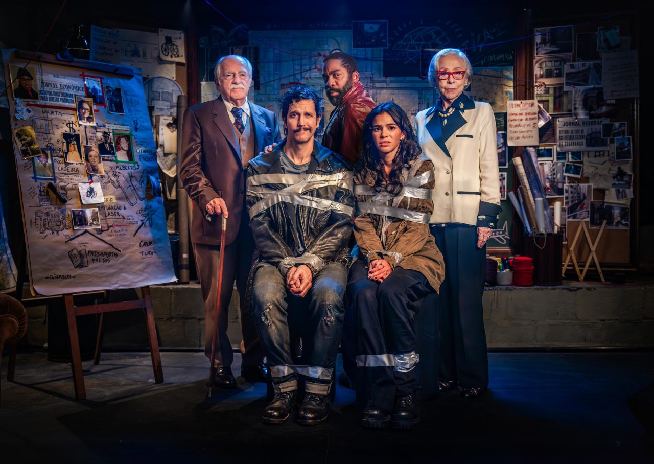 A group of six people, two taped together, poses in front of a wall covered in notes and photos.