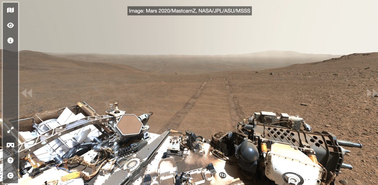 MARS 2020 Rover: Blick auf die staubige, helle Marsoberfläche mit Spuren und Gebirgen im Hintergrund.