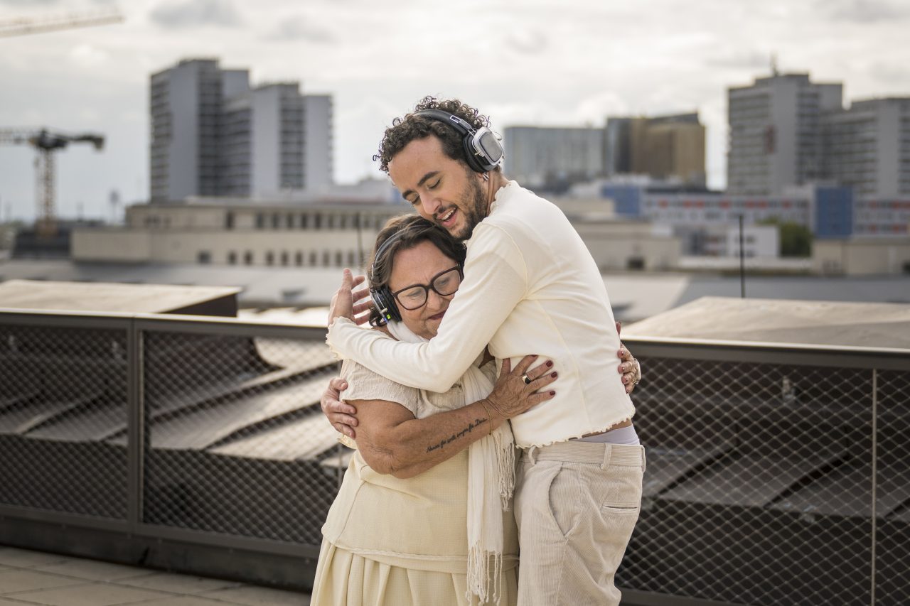 A man and an older woman lovingly embrace on a rooftop terrace, both wearing headphones.