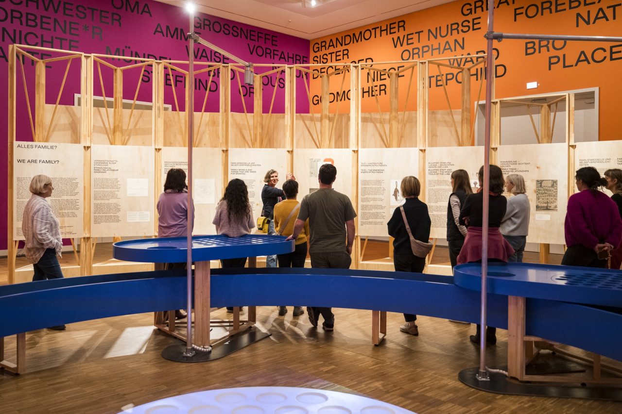 Besucher in einem Ausstellungsraum lesen informative Tafeln vor bunten Wänden und einer blauen Theke.