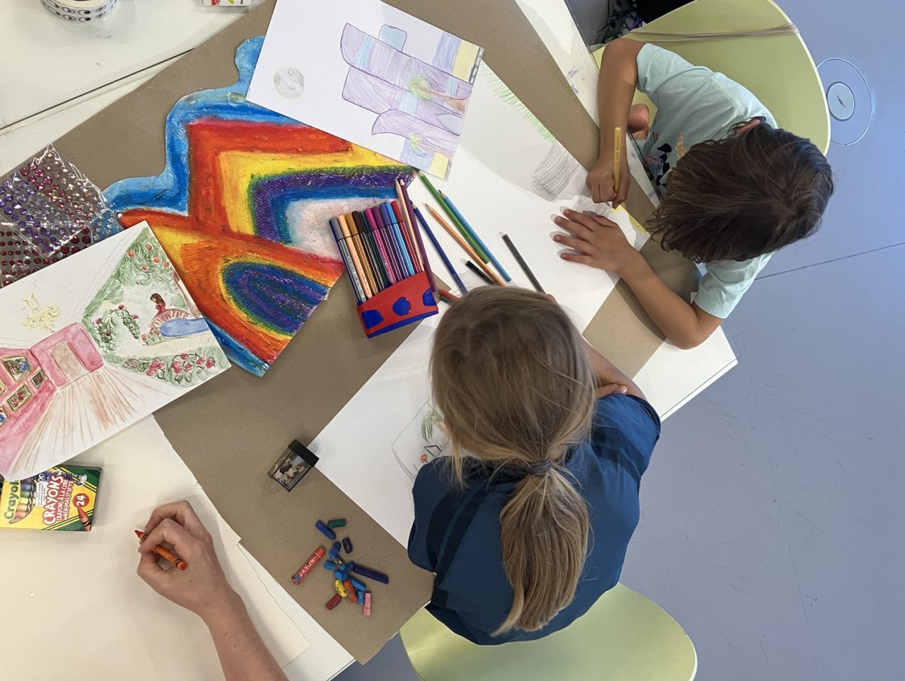 Children are drawing and coloring at a table, surrounded by pencils, paper, and colorful drawings.