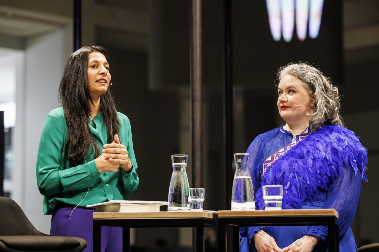 Zwei Frauen stehen am Tisch und unterhalten sich. Eine trägt eine grüne Bluse, die andere ein blaues Kleid.