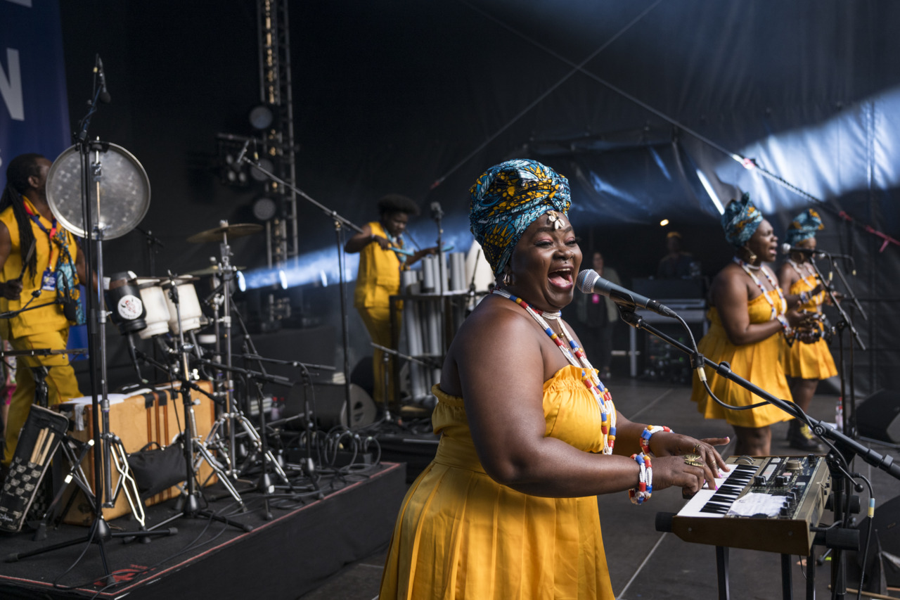 Stage with female musicians in yellow dresses singing and playing a keyboard, accompanied by a band.