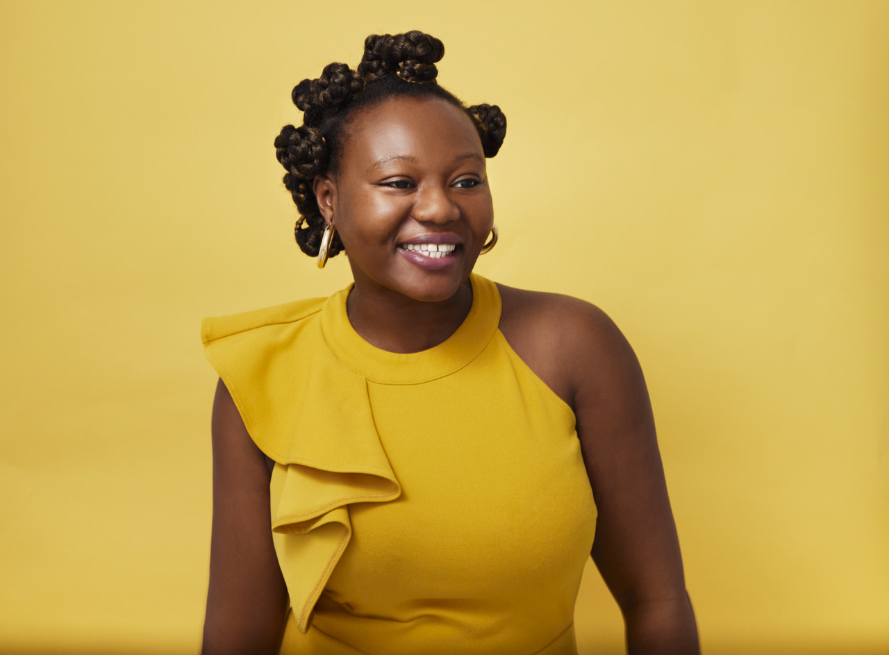 Smiling woman with curly hair in a yellow dress against a solid yellow background.