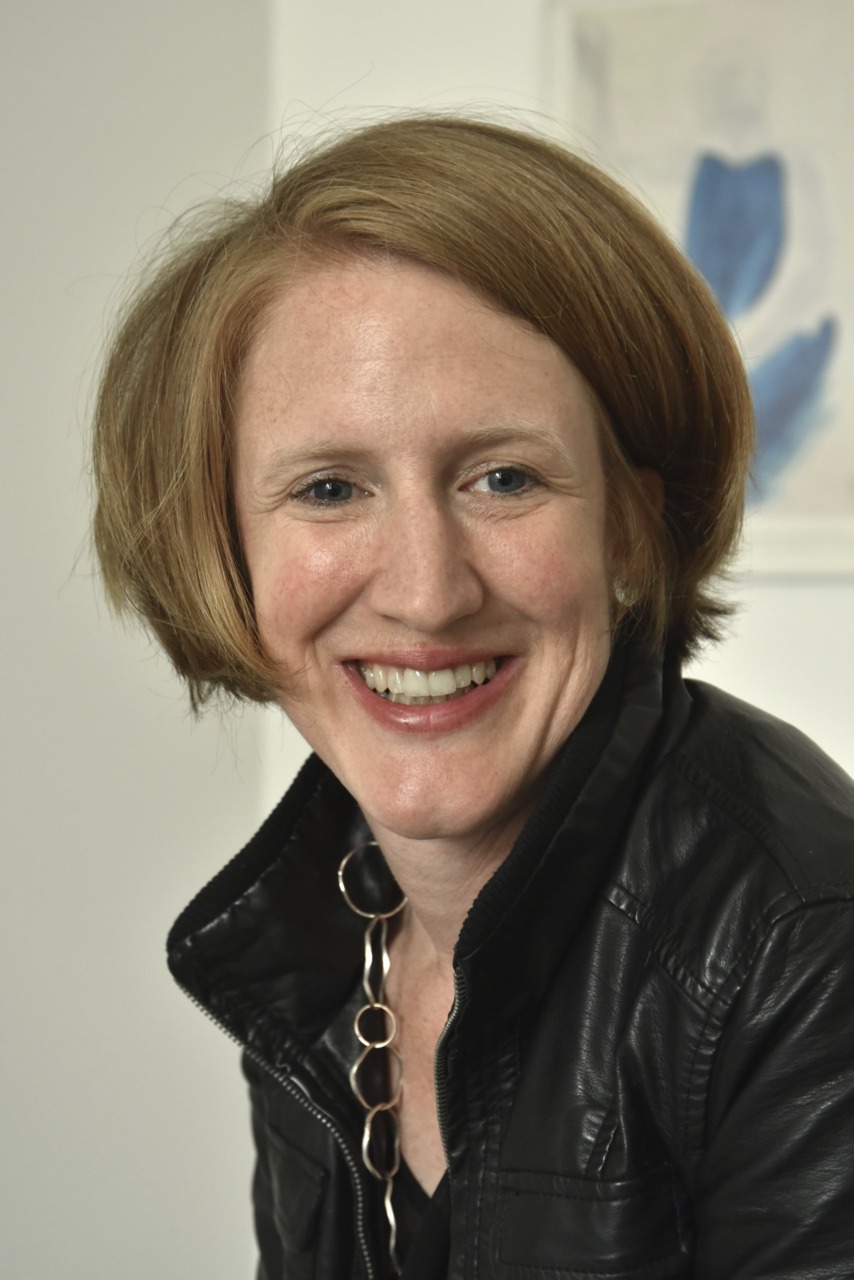 Smiling woman with short blonde hair, black jacket, and chain necklace, against a neutral background.