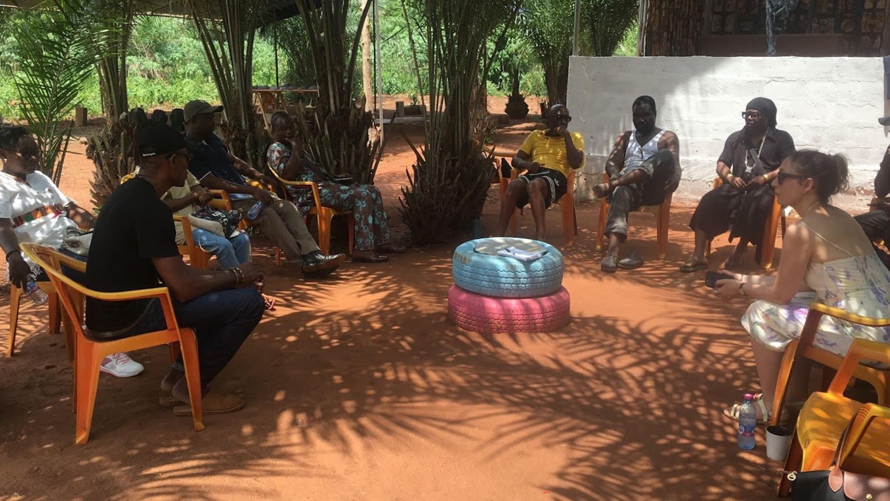 A group of people sits under trees on chairs in an informal outdoor meeting.